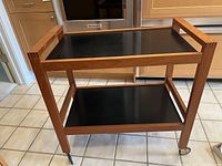 Front view of two-tier wood trolley with black shelves on tiled floor
