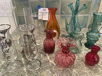 Wide view of the collection of art glass vases and cruets showing variety in colors and styles including colors such as red, pink, green, and clear glass with floral overlays and ruffle trims.