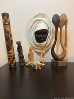 Wide angle photo showing all items including wooden rainstick, leather hanging mask, multiple carved figures, and large wooden abstract sculpture.