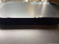 Front view of black Samsung Blu-ray DVD player on black surface, showing full width and top surface with some reflections.