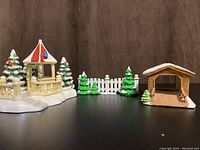 Village gazebo with red roof, snow-topped trees, and white picket fence with green snow-covered trees next to brown covered bridge