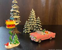 Several snow-covered Christmas trees, a yellow treehouse on a tree with a small snowy base and a red bird, and a pink convertible car decorated with Christmas items, all set against a dark background.