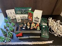 Lot arranged on table showing boxed lamp posts, street lamps, stone fencing, benches, snow covered trees, Santa and Mrs. Claus figurines, and red truck figurine