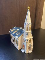 Front and side view of white chapel with blue roof and steeple with cross, showing gold arched windows and snow details.