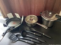Photo showing a 9-inch T-fal frying pan, 12-inch The Rock frying pan, stainless steel saucepan with lid, pressure cooker, and five black cooking utensils.