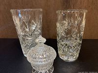 Front view of two crystal vases and one crystal candy dish with lid on brown background.