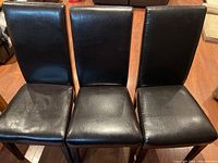 Three black leather dining chairs with wood legs photographed from a slight angle showing seat and back, placed on wooden floor.