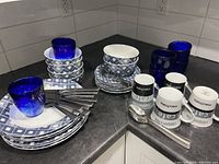Photo showing stacked patterned dinner plates, bowls, blue juice glasses, cappuccino mugs with text, and stainless steel utensils including knives, spoons, and forks.