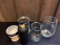 Five assorted flower vases including one black chalkboard finish vase with string detail and four clear/white glass vases on black surface, front view.