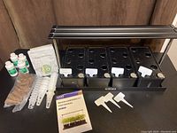 Full kit laid out showing black planter, LED light, plant labels, seed pods, liquid food bottles, soil medium packets, and user manual