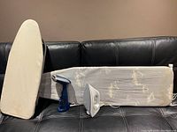 Standing ironing board and countertop ironing board placed on black leather couch alongside blue Conair steam press and white Tefal UltraGlide iron.