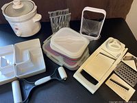 Photo showing small white slow cooker with glass lid, stainless steel pastry roller, white ceramic divided condiment server, collapsible storage containers with lids, and a white plastic mandolin slicer with attachments laid out on a black surface.