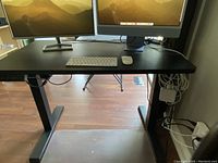 Front view of the desk showing dual monitors, keyboard, and mouse on the black desktop surface with T-shaped legs.