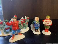 Four Snow Village figurines consisting of two wreath displays, a small elderly figure, and a boy holding a wreath, all on white bases with Christmas colors.