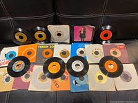 Photo showing 17 vintage 45 RPM vinyl records spread out with visible paper sleeves, showing variety of labels and wear on sleeves and records.