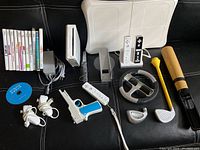 Full view of Nintendo Wii system components including console, controllers, balance board, sensor bar, and games laid out on black surface.