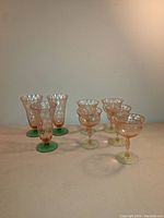 Full front view showing all 8 glasses: 3 taller parfait glasses with deep green foot and 5 shorter goblets with pale green foot, both with pink body and diamond pattern.