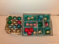 29 assorted vintage plastic Christmas ornaments displayed in original cardboard sectioned packaging with green tissue paper, featuring colors like green, red, gold, turquoise, and blue.