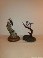 Front view of woodpecker ceramic figurine on wooden base and blue jay painted wood figurine on branch-like base.
