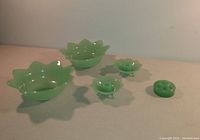 Five green jade-ite glass pieces: two lotus console bowls, two lotus candlestick holders, one flower frog displayed on flat surface.