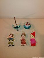 Front view of six vintage Christmas ornaments including dancing elf, felt elf, plastic angel, glass candle holder, crackled glass globe, and blue glass ball