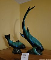 Photo showing two Blue Mountain Pottery fish sculptures side by side on a wooden surface against a yellow wall; the fish in the foreground has a long upward curved tail.