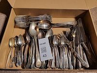 Wide view of a box containing multiple pieces of antique flatware including forks, spoons, and knives with tarnished silver plating.