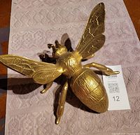 Top angle view of gold coloured metal bee resting on patterned mat showing wing detail and body texture.