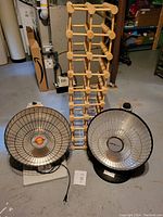 Photo showing two heat lamps placed side by side in front of a foldable wooden wine rack on a basement floor.