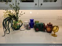 Full view of ceramic and glass decorative items on white counter including teapot, vases, goblet, and artificial plant.