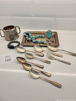 Photo showing silverware including teaspoons, a fork, a small silver handled cup, turquoise necklace and bracelet, and green bracelet on a white counter with lot number card.
