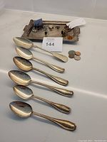 View of six silver spoons laid on a white surface alongside various rings, earrings, coins, and an antique tray in the background.