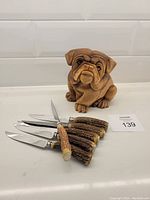 Photo showing all five cutting tools with horn handles and metal end caps, with one horn handle missing its metal end cap, and the wood carved bulldog figurine beside them on a white surface.