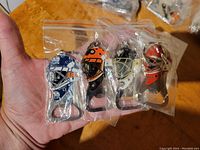Hand holding four NHL helmet bottle opener key chains individually packaged in clear plastic bags showing different team helmet designs in blue/white, orange/black, black/gold, and red/white