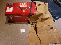 Red metal toolbox closed next to tan leather shop apron folded beside it showing overall size and condition
