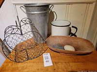 Photo showing all items in the lot on a wooden surface: large galvanized metal vase, heart-shaped wire basket, large white enamel mug, wooden bowl with stones.