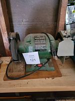 Front view of the green 6 inch bench grinder on wooden base with attached power cord and plug, labeled as lot 121.