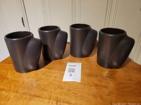 Four chocolate brown Beauceware ceramic mugs with sculptural handles shown from one angled side.