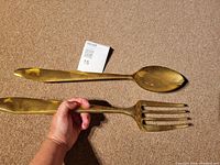 Two large gold color heavy metal kitchen utensils, a spoon and a fork, side by side on carpet next to a size reference item.