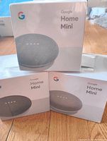 Three sealed Google Home Mini boxes with one showing front with speaker image and branding.