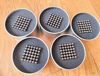 5 round metal padded cases each containing 216 black magnetic balls arranged in a square grid formation.