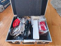 Open black aluminum case showing neatly arranged tattoo machines, power supply, cables, red ink cap holder and plastic bags of accessories.