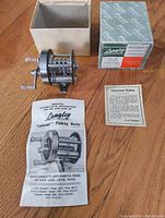 Angled shot of the Langley Plugcast fishing reel with original box and printed materials laid out on wood surface.