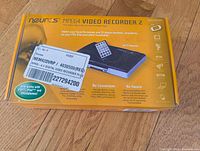 Neuros MPEG4 Video Recorder 2 sealed in original yellow box placed on wooden floor, showing front and top sides with product image and key features.