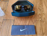 Nike Charger sunglasses in hard black protective case with cleaning cloth and Nike branded label shown on floor background.