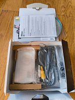 Open box showing Nikon Coolpix S3200 camera wrapped in protective plastic alongside manuals, USB cable, AV cable, and battery charger inside original packaging.