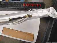 Full view of the Onikiri sword in white sheath with brass fittings lying next to original black and red Onikiri box on a white table.