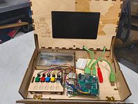Open wooden box showing inside components: circuit board, colored buttons, screen, wiring, and breadboard inside a laser-cut wooden case.