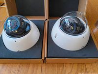 Two white dome security cameras with clear domes on bases inside original packaging boxes.