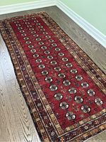 Full view of the Afghani Bokhara runner showing the main red field with rows of geometric medallion motifs and the multi-banded border in earth tones on wooden floor.
